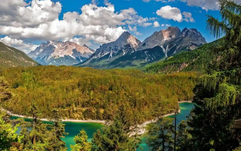 Panoramic alpine valley with turquoise lake, dense evergreen forest, and snow-dusted peaks beneath bright clouds—featured image for spiritual poem about nature's beauty